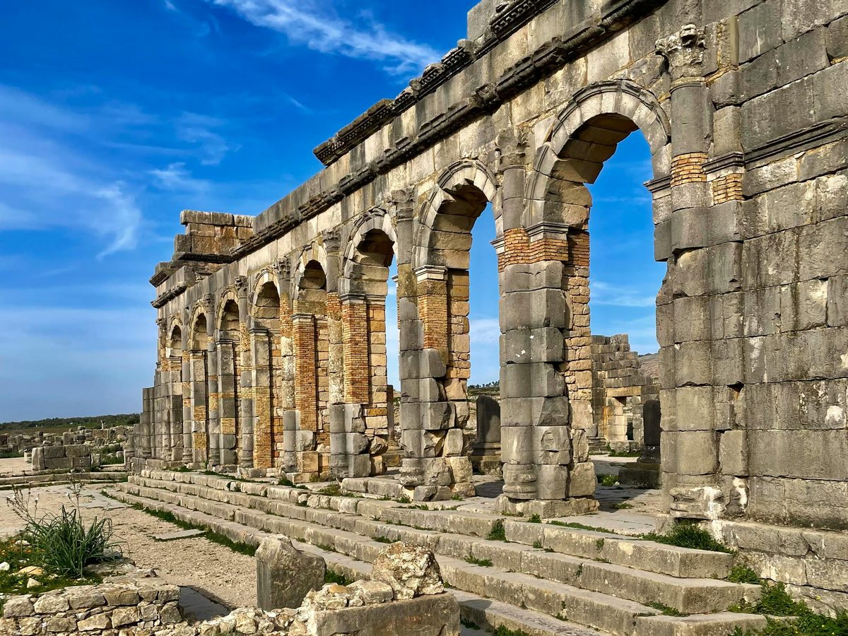 The Roman basilica of Volubilis — a row of stone arches and brick columns standing against a vivid blue sky